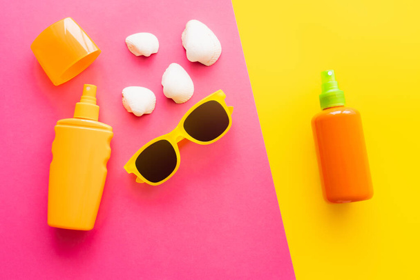 Sonnencreme in Flaschen in der Nähe von Sonnenbrille und Muscheln auf rosa und gelben Hintergrund  - Foto, Bild