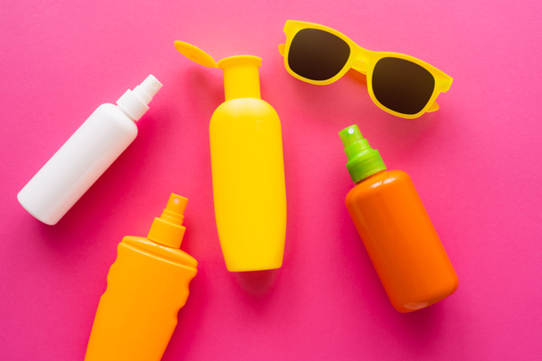 Draufsicht auf Flaschen mit Sonnenschutzmitteln in der Nähe von Sonnenbrillen auf rosa Oberfläche  - Foto, Bild