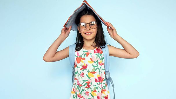 Studio portrait of pretty student girl putting and holding academic book on her head and looking at the camera, wearing colorful dress, eyeglasses, and backpack posing on blue background. - Foto, imagen