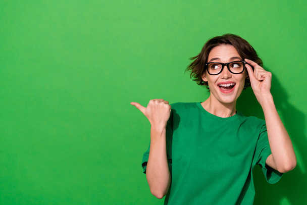Фото чарівної смішної жінки в повсякденних футболках окулярах, спрямованих на великий палець, що виглядає порожнім простором ізольований зелений колір фону
. - Фото, зображення