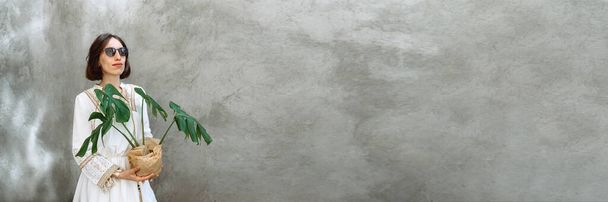 Baner γίνεται με φωτογραφία Το κορίτσι σε όμορφο λευκό φόρεμα boho στυλ κατέχει στο φυτό γλάστρα Monstera με γοητευτικό πράσινα φύλλα σε ένα φόντο της μόδας τσιμεντένιο τοίχο. Γυναίκα με γυαλιά ηλίου. Αντιγραφή χώρου - Φωτογραφία, εικόνα