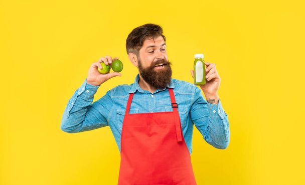 Happy man in apron looking at juice bottle holding limes yellow background, juice barman. - Foto, imagen