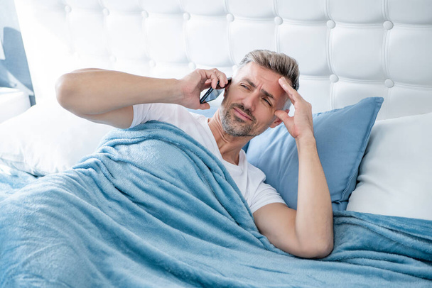 sleepy mature man in bed speaking on phone. headache. - Zdjęcie, obraz