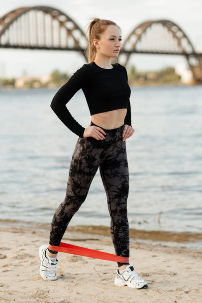 Sports girl performs a workout with fitness rubber bands on the river bank - Photo, Image