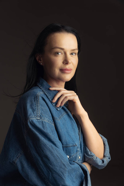 Elegance. Portrait of attractive forty year old woman in blue denim shirt posing isolated on dark background. Concept of live portrait, beauty, fashion. Model with long black hair. Copy space for ad - Foto, immagini