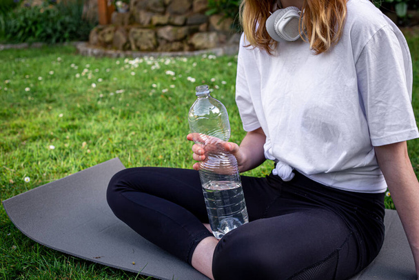 Uma jovem mulher em um tapete de água potável de uma garrafa na natureza, o conceito de praticar esportes, treinamento e fitness. - Foto, Imagem