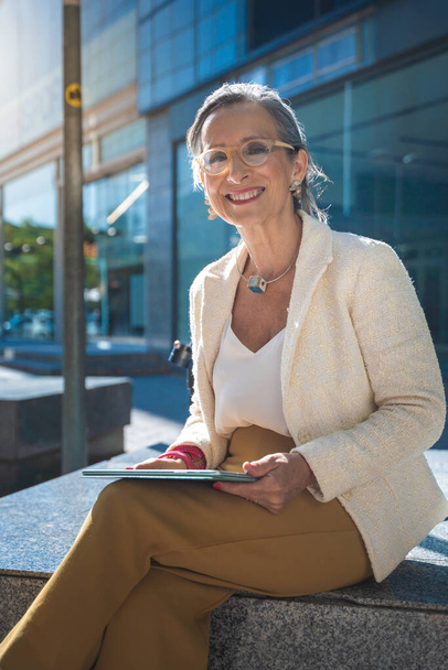 Happy attractive business woman middle-aged looking to camera smiling with technology tablet in front of a modern building. High quality photo - Фото, изображение