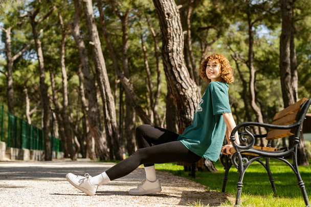 Happy redhead woman wearing green tee standing on city park, outdoor concentrating on her workout, leaning on a park bench, doing triceps exercise. - Photo, Image