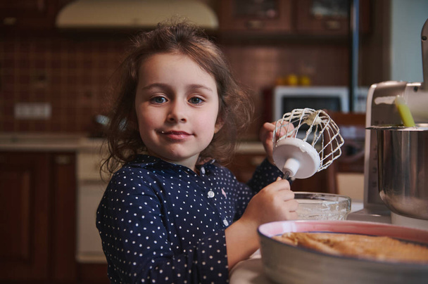 Retrato de uma linda criança caucasiana, menina olhando para a câmera enquanto lambe batedor com sobras de chantilly em tigela de aço na cozinha doméstica - Foto, Imagem