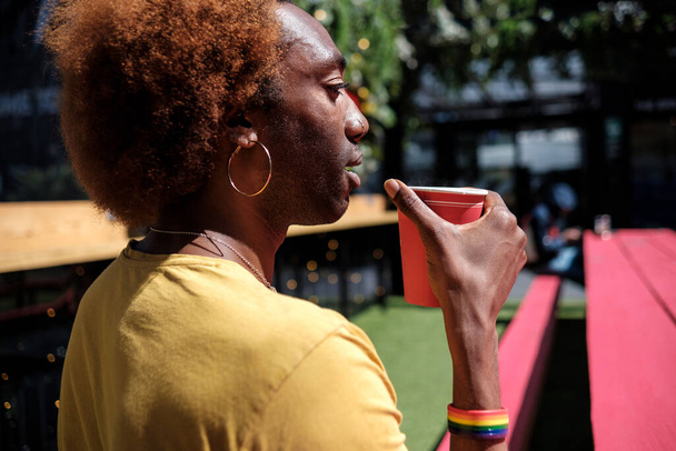 Νεαρό μη δυαδικό άτομο που πίνει καφέ καθισμένος σε ένα πολύχρωμο κόκκινο παγκάκι. Φοράει ένα κίτρινο μπλουζάκι. Σγουρά ξανθά μαλλιά. - Φωτογραφία, εικόνα