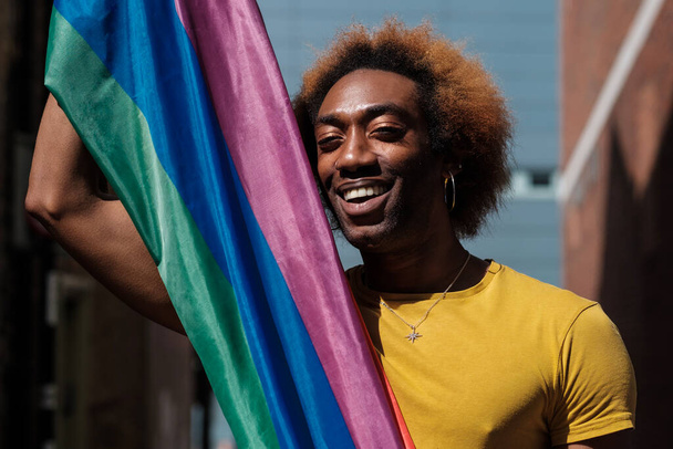 Retrato de una persona sonriente no binaria sosteniendo una bandera lgbtq y mirando a la cámara. Lleva camisa amarilla. Cabello rubio rizado. - Foto, Imagen