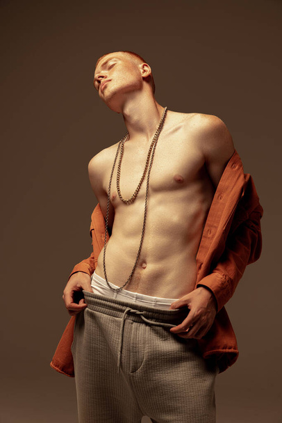 Portrait of young stylish red-haired man posing in unbuttoned oramge shirt, wearing trendy necklace isolated on grey studio background. Concept of fashion, style, body aesthetics, beauty, mens health - Photo, Image