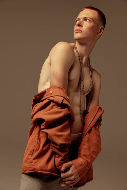 Portrait d'un beau jeune homme roux posant isolé sur fond de studio brun. Style masculin, accessoires. Concept de mode, style, esthétique corporelle, beauté, santé masculine, fitness - Photo, image