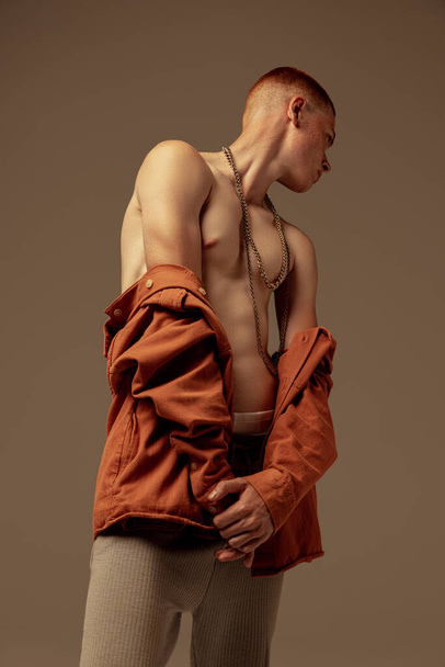 Портрет красивого молодого рыжего мужчины, позирующего изолированно на фоне коричневой студии. Мышечная спортивная красота тела. О моде, стиле, эстетике тела, красоте, здоровье, фитнесе - Фото, изображение