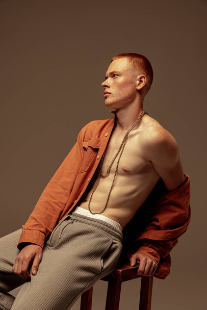 Portrait of young stylish red-haired man sitting on chair, posing in orange shirt isolated over brown studio background. Concept of fashion, style, body aesthetics, beauty, mens health - Foto, Imagem