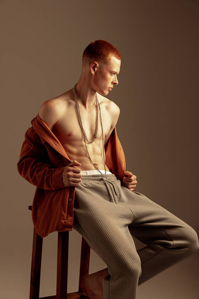 Портрет молодого стильного рыжего мужчины, сидящего на стуле, позирующего в оранжевой рубашке на коричневом фоне. Облегчение формы тела. Концепция моды, стиля, эстетики тела, красоты, мужского здоровья - Фото, изображение