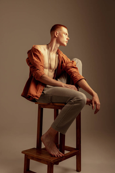 Portrait of young stylish red-haired boy sitting on chair, posing isolated over brown studio background. Body art. Concept of fashion, style, body aesthetics, beauty, mens health, fitness - Foto, afbeelding