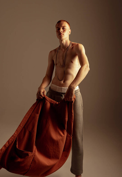 Портрет красивого рыжеволосого молодого человека, позирующего без рубашки на фоне коричневой студии. Мужские аксессуары. Боди-арт Концепция моды, стиля, эстетики тела, красоты, мужского здоровья - Фото, изображение
