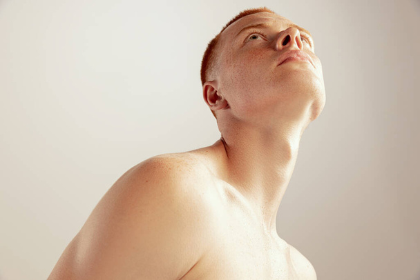 Portret van een jonge roodharige man die shirtloos poseert, naar boven kijkend geïsoleerd over grijze studio-achtergrond. Gesproeid lichaam. Concept van gezondheid, levensstijl, schoonheid, lichaams- en huidverzorging, mode - Foto, afbeelding