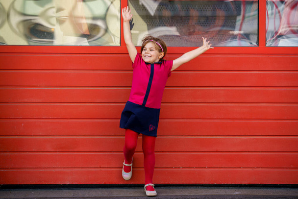 Портрет счастливой улыбающейся девочки в красной одежде на улице. Маленький ребенок со светлыми волосами, смотрящий и улыбающийся в камеру. Счастливый здоровый ребенок получает удовольствие от активного отдыха и игр - Фото, изображение