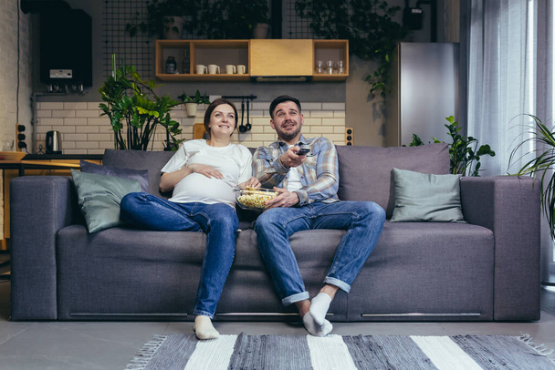 Home cinema. Young family. Pregnant woman and man sitting on sofa at home, watching TV, movie, eating popcorn. Have a good time together - 写真・画像