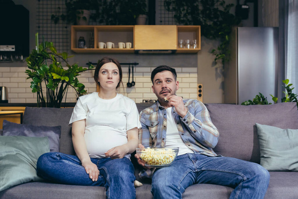 Ev sineması. Genç bir aile. Hamile kadın ve erkek evdeki kanepede oturmuş televizyon, film izliyor, patlamış mısır yiyorlar. Birlikte iyi vakit geçirin. - Fotoğraf, Görsel