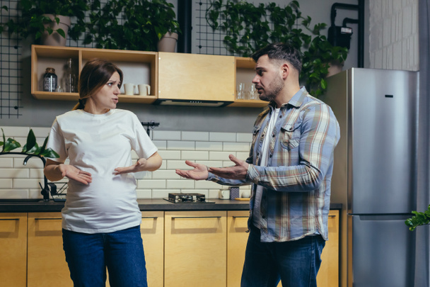 Family conflict. Young family. A pregnant woman and a man quarrel at home, shouting. They stand at home in the kitchen - Foto, Imagen