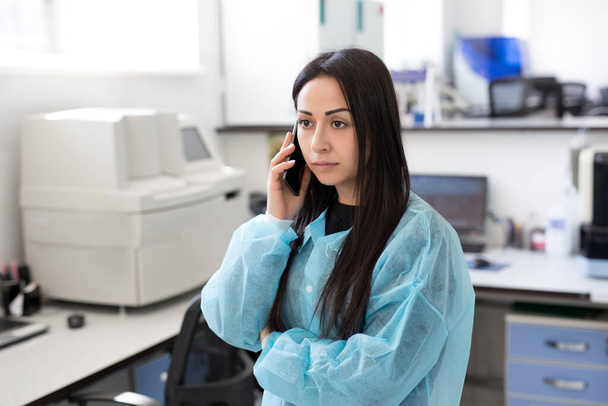 Attraktive Labormitarbeiterin mit Handy an ihrem Arbeitsplatz im Labor. Wissenschaftler diskutiert Analyseergebnisse mit Hilfe von Handy - Foto, Bild