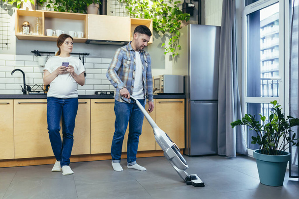 The young man helps his wife around the house. vacuum the apartment, house. The pregnant wife is standing next to her husband, drinking coffee, resting - Photo, image