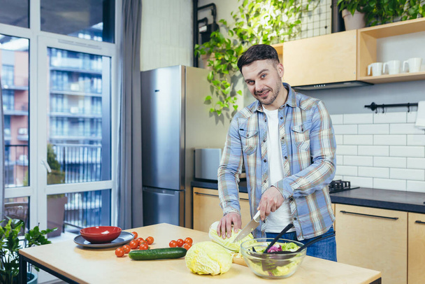 Young handsome man prepares healthy and tasty food in the kitchen at home. Meal and dinner. chops vegetables and fruits. He looks at the camera, smiles - Zdjęcie, obraz