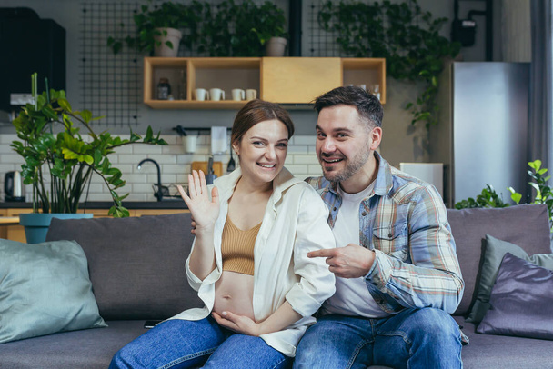 Young family sitting on sofa at home. They talk on a video call. Pregnant woman and man. Woman waving her hand at the camera, smiling. The man points a finger at her belly. Happy - Фото, зображення