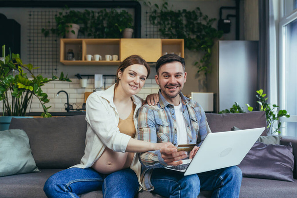 Online shopping. Young family, pregnant woman and man make an online purchase order from a laptop, holding a credit card. Sitting at home on the couch. Very happy, satisfied. They look at the camera and smile - Photo, Image