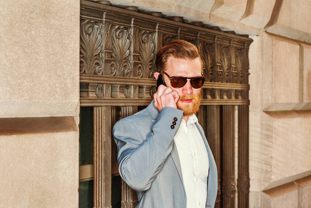 Молодой американский бизнесмен с бородой в голубой куртке, белой майке, солнечных очках, стоит напротив старинной стены на улице в Нью-Йорке, разговаривает по мобильному телефону. - Фото, изображение