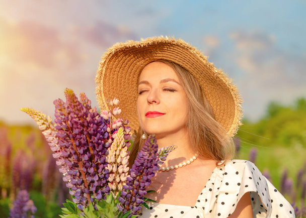 a young woman with a bouquet of lupines in a polka dot dress and a straw hat walks on the lawn with lupin at sunset - Photo, Image