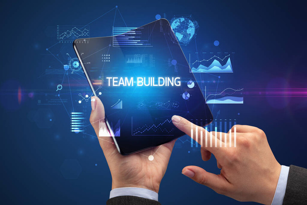 Businessman holding a foldable smartphone with TEAM BUILDING inscription, successful business concept - Photo, Image
