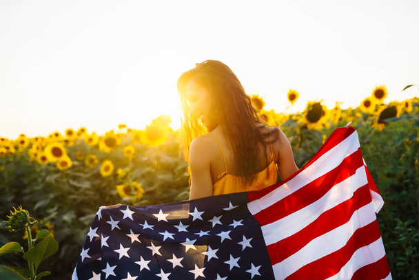 Beautiful girl in hat with the American flag in a sunflower field. 4th of July. Fourth of July. Freedom. Sunset light The girl smiles. Beautiful sunset. Independence Day. Patriotic holiday. - Photo, image