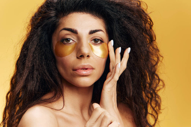 Curly pretty Latin woman in putting gold hydrogel mask on face for under-eye skin repair posing isolated over yellow background. Cosmetic product ad Natural beauty concept Studio closeup portrait - Photo, Image