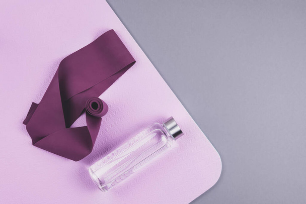 Draufsicht auf kastanienbraunes Expander-Gummiband, wiederverwendbare Glas-Wasserflasche auf rosa Yogamatte vor neutralem grauen Hintergrund. - Foto, Bild