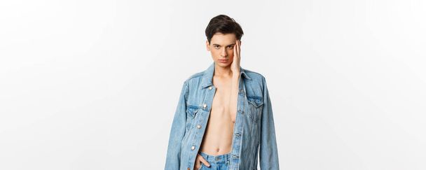 Handsome and sassy gay man wearing denim jacket on bare torso, touching his face and looking confident at camera, standing over white background. - Foto, afbeelding