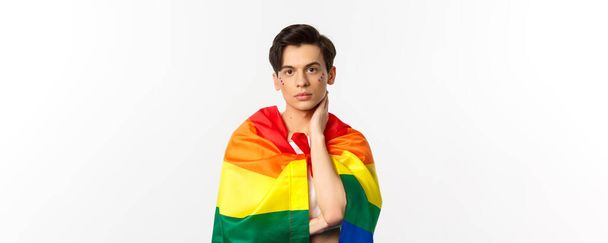 Waist-up shot of beautiful gay man with glitter on face, wrap himself with rainbow lgbt flag and touching face gently, looking at camera, white background. - Photo, Image