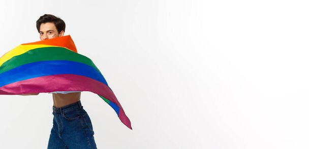 Derechos humanos y concepto de comunidad lgbtq. Joven queer con brillo en la cara, ondeando bandera lgbtq con orgullo, de pie sobre fondo blanco. - Foto, imagen