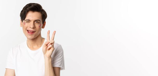 People, lgbtq community and lifestyle concept. Happy and cute gay man with glitter on face, showing peace sign and smiling, celebrating pride holiday, white background. - Photo, Image