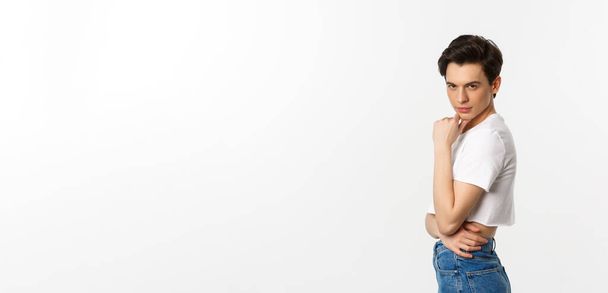 Lgbtq and pride concept. Side view of attractive androgynous man in crop top looking sassy at camera, standing over white background. - Foto, afbeelding