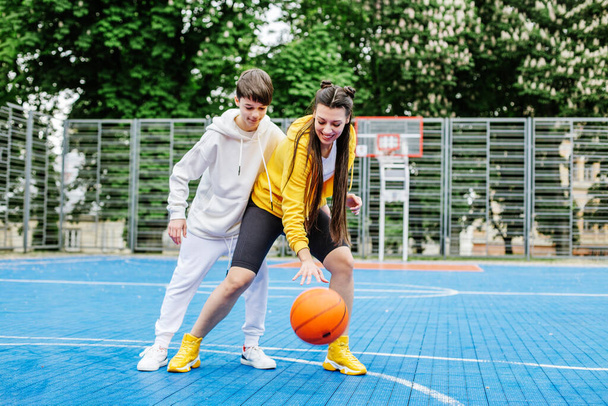 Κορίτσι και ο μικρότερος αδελφός της, έφηβος, παίζουν μπάσκετ στο γήπεδο του μοντέρνου μπάσκετ κάτω από τον ουρανό. Έννοια του αθλητισμού, χόμπι και υγιεινό τρόπο ζωής. - Φωτογραφία, εικόνα