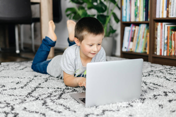 Ο γοητευτικός σχολιαρόπαιδο ξαπλώνει στο πάτωμα και παρακολουθεί μαθήματα ή συζητήσεις στο διαδίκτυο με φίλους. Online μάθηση και χρήση gadget από τα παιδιά - Φωτογραφία, εικόνα