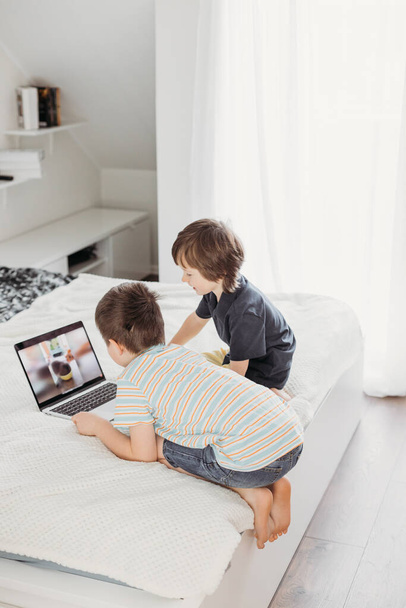 自宅での距離学習。子供たちはデジタルタブレットやラップトップを使用して学びます。少年たちはベッドの上で何かを見ている. - 写真・画像