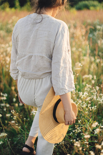 Elegante mujer boho con sombrero de paja caminando entre flores silvestres en la cálida luz del atardecer. Delicias de verano y viajes. Joven hembra en tela de lino rústico relajándose en el prado de verano. Momento atmosférico - Foto, imagen