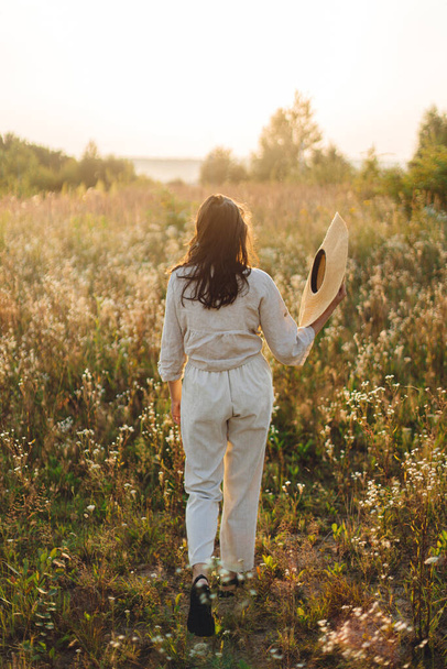 Stylische Boho-Frau mit Strohhut, die zwischen Wildblumen im warmen Sonnenuntergang wandelt. Sommerfreude und Reisen. Junges Weibchen in rustikalem Leinentuch entspannt auf der Sommerwiese. Atmosphärischer Moment - Foto, Bild