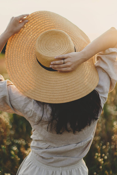Mulher boho elegante com chapéu de palha posando entre flores silvestres na luz do pôr do sol, visão traseira. Delícia de verão e viagens. Jovem fêmea despreocupada em pano de linho rústico relaxante no prado de verão. Atmosférico - Foto, Imagem