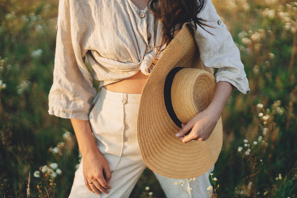 Young carefree female in rustic linen cloth close up relaxing in summer meadow. Summer delight and travel. Stylish boho woman with straw hat posing among wildflowers in sunset light - Foto, afbeelding